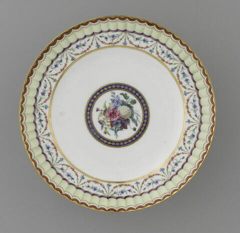 Assiette, d'une paire (R 758)
Sèvres, vers 1793; marque au revers
Au fond, un bouquet de fleurs, entouré d'un cercle bleu ponctué d'or. Au marli, guirlande de soses et de bleuets suspendue à un lerge ruban jaune bordé de rouge et or.