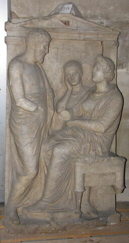 stèle ; Tirage d’une stèle funéraire attique
Le haut-relief représente un homme (Thraséos du dème de Perithoidai), debout de trois-quarts et vêtu d'un himation, serrant la main (dexiosis) d'une femme (Evandréia), assise sur un tabouret, vêtue d'un chiton et d'un himation. Entre eux, à l'arrière-plan apparaît une petite fille (servante) la tête appuyée sur la main droite en signe d'affliction. Une inscription en grec, gravée sur l'architrave, indique les noms des personnages.
Le moulage ne présente ni les mains droites de la femme et de l'homme debout, ni le pied arrière du siège.