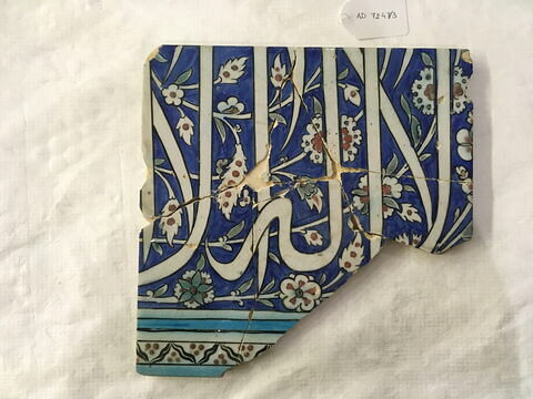 Carreau à inscription et rinceaux à rosettes et fleurettes sur fond bleu. Bordure à cintemani et lèvres de Bouddha